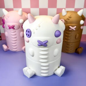 May include: Three whimsical, cylindrical containers in pink, white, and brown, each with a cartoonish monster design. They feature horns, button eyes, and a stitched mouth. The containers are on a purple surface with a pink and white checkered background.