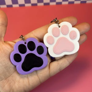 May include: Two paw-shaped keychains. One is lavender with black paw pads, the other is white with pink paw pads. Both have silver key rings attached. The keychains are held in a hand against a pink and purple checkered background.