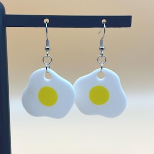 May include: A pair of novelty earrings shaped like fried eggs. Each earring features a white egg with a yellow yolk and a silver-colored hook. The earrings are displayed against a neutral background.