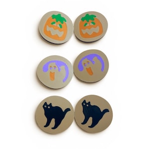 May include: Six circular beige tokens arranged in three pairs. The top pair features orange jack-o'-lanterns with green stems. The middle pair displays purple ghosts with orange mouths. The bottom pair shows black silhouettes of arched cats.