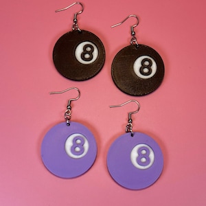May include: Four earrings, two brown and two purple, each featuring a number 8 ball design. The brown earrings have a textured surface, while the purple ones are smooth. All earrings have silver-colored hooks.