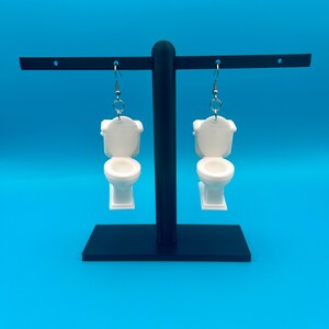 May include: White toilet-shaped earrings hanging from silver hooks. The earrings are displayed on a black stand against a bright blue background. The earrings are a novelty item.