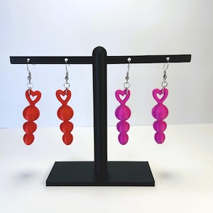 May include: A black earring display stand holding four pairs of earrings. Two pairs are red, and two are pink. Each earring has a heart-shaped top and three round beads hanging below.