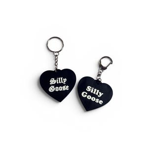 May include: Two black heart-shaped keychains with silver-toned metal rings and chains. Each keychain features the text "Silly Goose" printed in white, with one using a gothic-style font and the other using a standard sans-serif typeface.