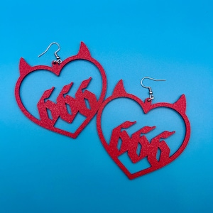 May include: A pair of red heart-shaped earrings with devil horns and the number 666 inside. The earrings are made of a glittery material and have silver-colored fish hook ear wires. The background is a solid blue.