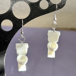 May include: A pair of novelty earrings featuring miniature white toilet designs. Each earring has a silver-colored fish hook and a small ring connecting the toilet charm. The earrings are displayed against a soft purple background.