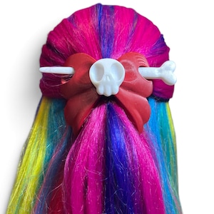 May include: A hair accessory featuring a red bow with a white skull center and a white bone-shaped hair stick. The accessory is secured in vibrant, multi-colored hair with streaks of pink, blue, yellow, and purple throughout the strands.