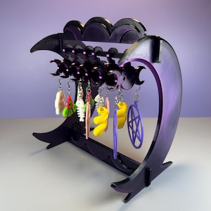 May include: A purple earring display stand with a crescent moon design. The stand holds various colorful earrings, including shapes like a fish, a spiral, and a pentagram. The stand is made of a dark purple material.