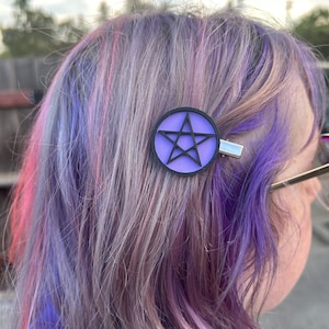 May include: A purple and black pentagram hair clip. The hair clip is round with a black pentagram design on a purple background. The clip is attached to multi-colored hair with shades of purple, pink, and gray.