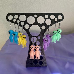 May include: A black earring display stand holds several pairs of alien-shaped earrings. The earrings are in various colors, including blue, yellow, pink, purple, and green. The stand has a unique, perforated design.