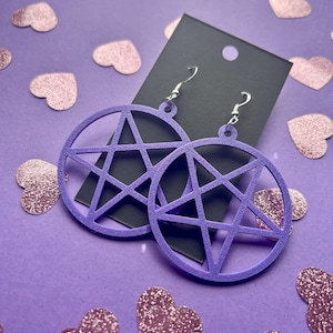 May include: A pair of lavender pentagram earrings with silver hooks. The earrings are displayed on a black card against a purple background scattered with glittery heart shapes. The earrings are a circular design with a five-pointed star inside.