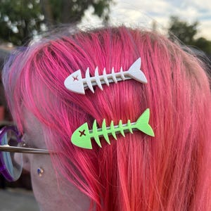 May include: Two fish bone hair clips. One clip is white, the other is neon green. Both clips are in the shape of a fish skeleton with an "X" on the head. The clips are in pink and red hair.