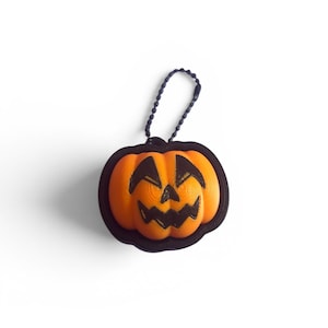 May include: A bright orange pumpkin-shaped charm with a black carved face featuring triangular eyes and a jagged mouth. The pumpkin is encased in a black border and includes a black ball chain attachment for hanging, creating a small accessory.