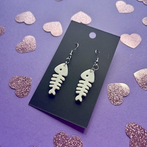 May include: A pair of white fishbone earrings with silver-colored hooks are displayed on a black card. The earrings are set against a purple background scattered with pink glitter heart-shaped confetti.