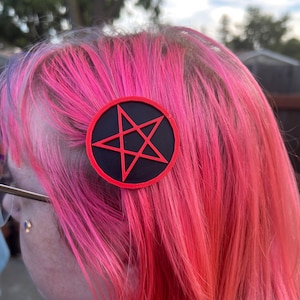 May include: A black and red pentagram hair clip is attached to vibrant pink and red hair. The hair clip is round with a black background and a red outline and star design. The hair has a gradient effect, transitioning from pink to red.