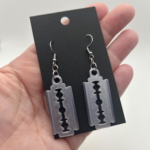 May include: A pair of silver-colored earrings shaped like razor blades. The earrings are hanging from silver-colored hooks and are attached to a black card. The earrings have a detailed design with multiple holes.