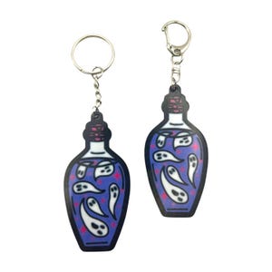 May include: Two keychains shaped like potion bottles. The bottles are dark blue with white ghost designs and pink star accents. Each keychain has a silver metal ring or clasp attached to the top.