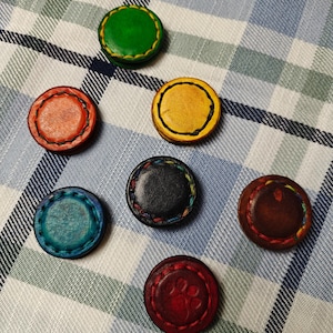 May include: Seven colorful, round leather coasters. The coasters are in shades of green, orange, yellow, teal, black, brown, and red. The red coaster has a paw print design. Each coaster has a stitched border.