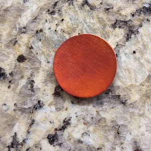 Peut inclure: Un dessous de verre rond en bois de couleur orange. Le dessous de verre est de couleur unie et a une surface lisse. Il est pos&eacute; sur un comptoir en granit.