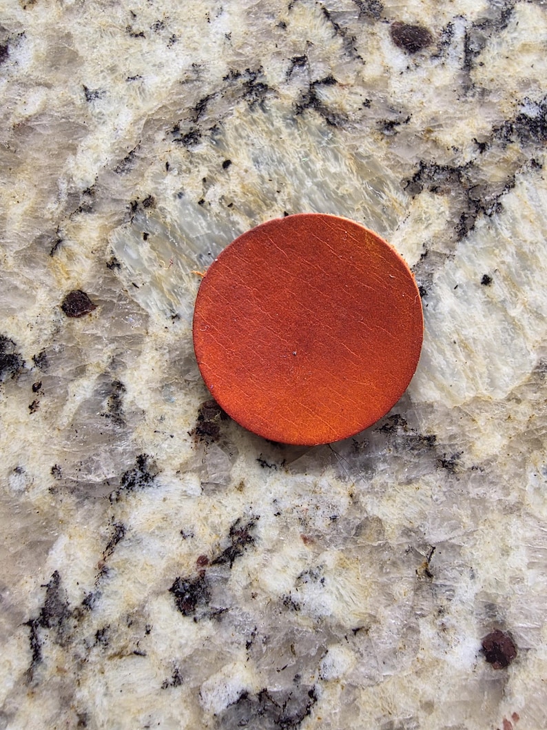 Peut inclure: Un dessous de verre rond en bois de couleur orange. Le dessous de verre est de couleur unie et a une surface lisse. Il est pos&eacute; sur un comptoir en granit.