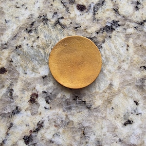 Peut inclure: Un petit objet rond de couleur or. L'objet est un cercle plein avec une surface l&eacute;g&egrave;rement textur&eacute;e. Le fond est une pierre de couleur claire avec des veines grises fonc&eacute;es et marron.