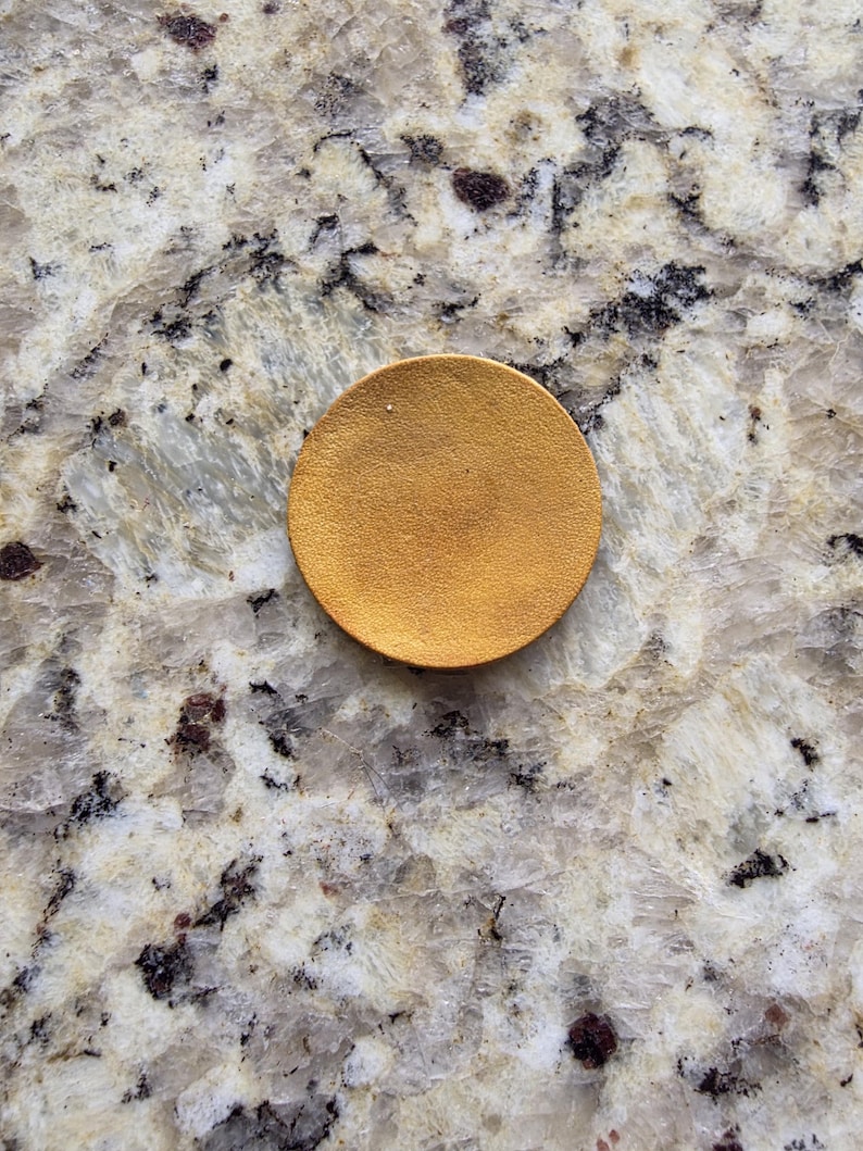 Peut inclure: Un petit objet rond de couleur or. L'objet est un cercle plein avec une surface l&eacute;g&egrave;rement textur&eacute;e. Le fond est une pierre de couleur claire avec des veines grises fonc&eacute;es et marron.