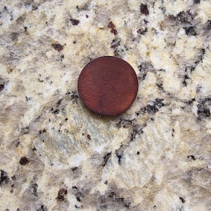 Peut inclure: Un petit objet rond, brun fonc&eacute;, possiblement un bouton en bois ou un &eacute;l&eacute;ment d&eacute;coratif, repose sur une surface tachet&eacute;e de couleur claire. La couleur de l'objet contraste avec l'arri&egrave;re-plan, soulignant sa forme et sa texture. L'image est un gros plan.