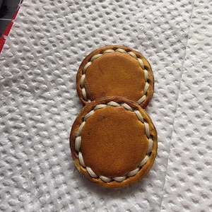 Peut inclure: Deux pi&egrave;ces rondes en cuir bronz&eacute; avec des coutures blanches sur les bords. Le cuir pr&eacute;sente un aspect l&eacute;g&egrave;rement vieilli. Elles sont pos&eacute;es sur une surface blanche textur&eacute;e.