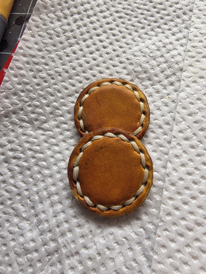 Peut inclure: Deux pi&egrave;ces rondes en cuir bronz&eacute; avec des coutures blanches sur les bords. Le cuir pr&eacute;sente un aspect l&eacute;g&egrave;rement vieilli. Elles sont pos&eacute;es sur une surface blanche textur&eacute;e.