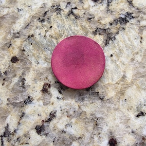 Peut inclure: Un objet rond de couleur magenta, possiblement un produit de maquillage, est pos&eacute; sur une surface claire et mouchet&eacute;e. L'objet est d'une couleur unie et uniforme avec un aspect l&eacute;g&egrave;rement textur&eacute;.