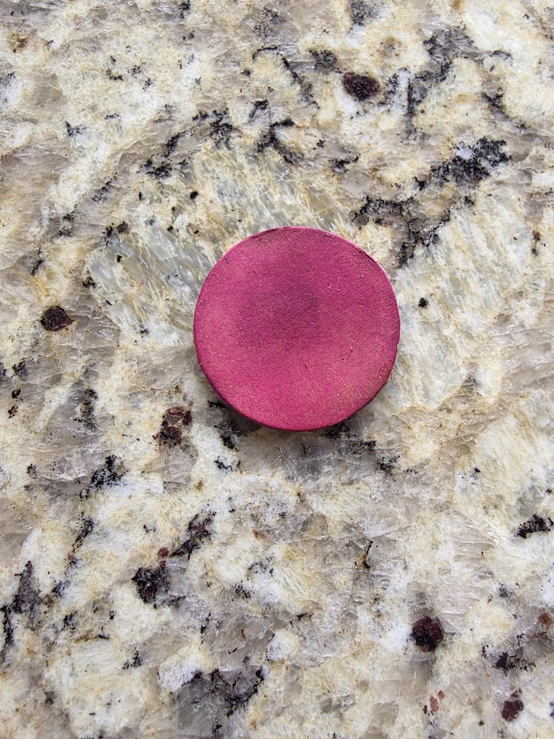 Peut inclure: Un objet rond de couleur magenta, possiblement un produit de maquillage, est pos&eacute; sur une surface claire et mouchet&eacute;e. L'objet est d'une couleur unie et uniforme avec un aspect l&eacute;g&egrave;rement textur&eacute;.