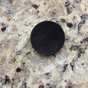 Peut inclure: Un petit dessous de verre rond en cuir noir. Le dessous de verre est circulaire et semble &ecirc;tre fait d'un mat&eacute;riau en cuir textur&eacute;. Le dessous de verre est pos&eacute; sur une surface claire et mouchet&eacute;e.
