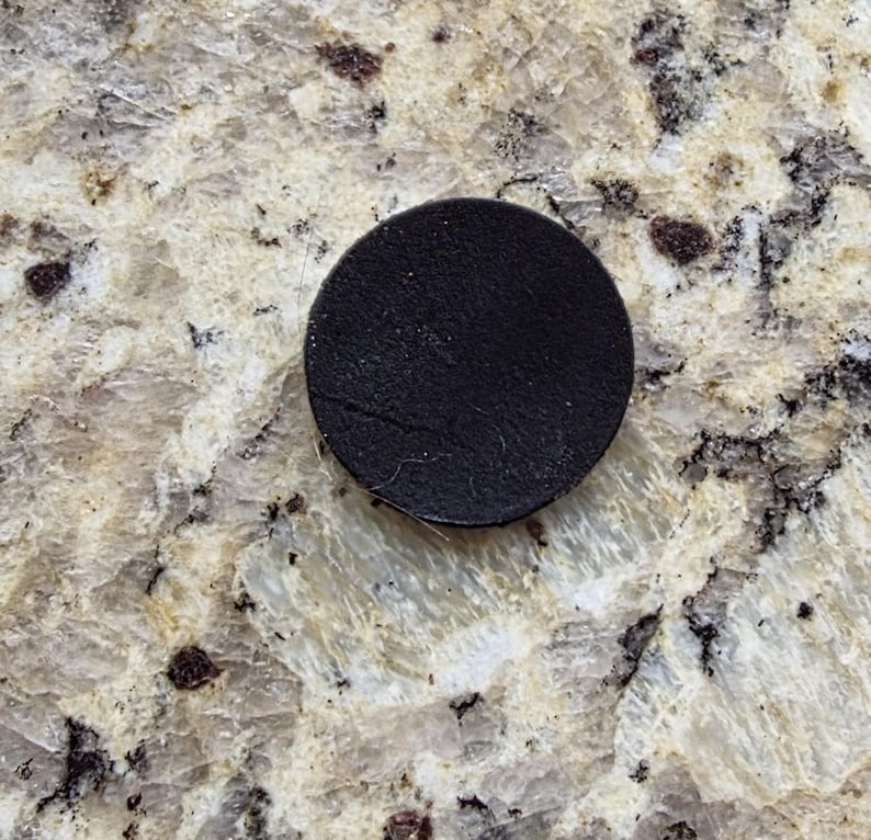 Peut inclure: Un petit dessous de verre rond en cuir noir. Le dessous de verre est circulaire et semble &ecirc;tre fait d'un mat&eacute;riau en cuir textur&eacute;. Le dessous de verre est pos&eacute; sur une surface claire et mouchet&eacute;e.