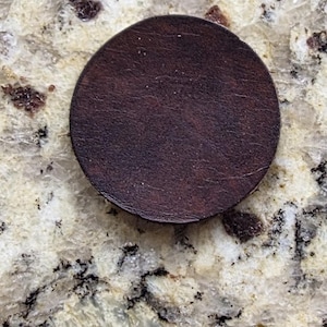 Peut inclure: Un dessous de verre circulaire en cuir marron fonc&eacute;. Le dessous de verre est de couleur unie avec une surface l&eacute;g&egrave;rement textur&eacute;e. Le dessous de verre est pos&eacute; sur un comptoir en granit clair et mouchet&eacute;.