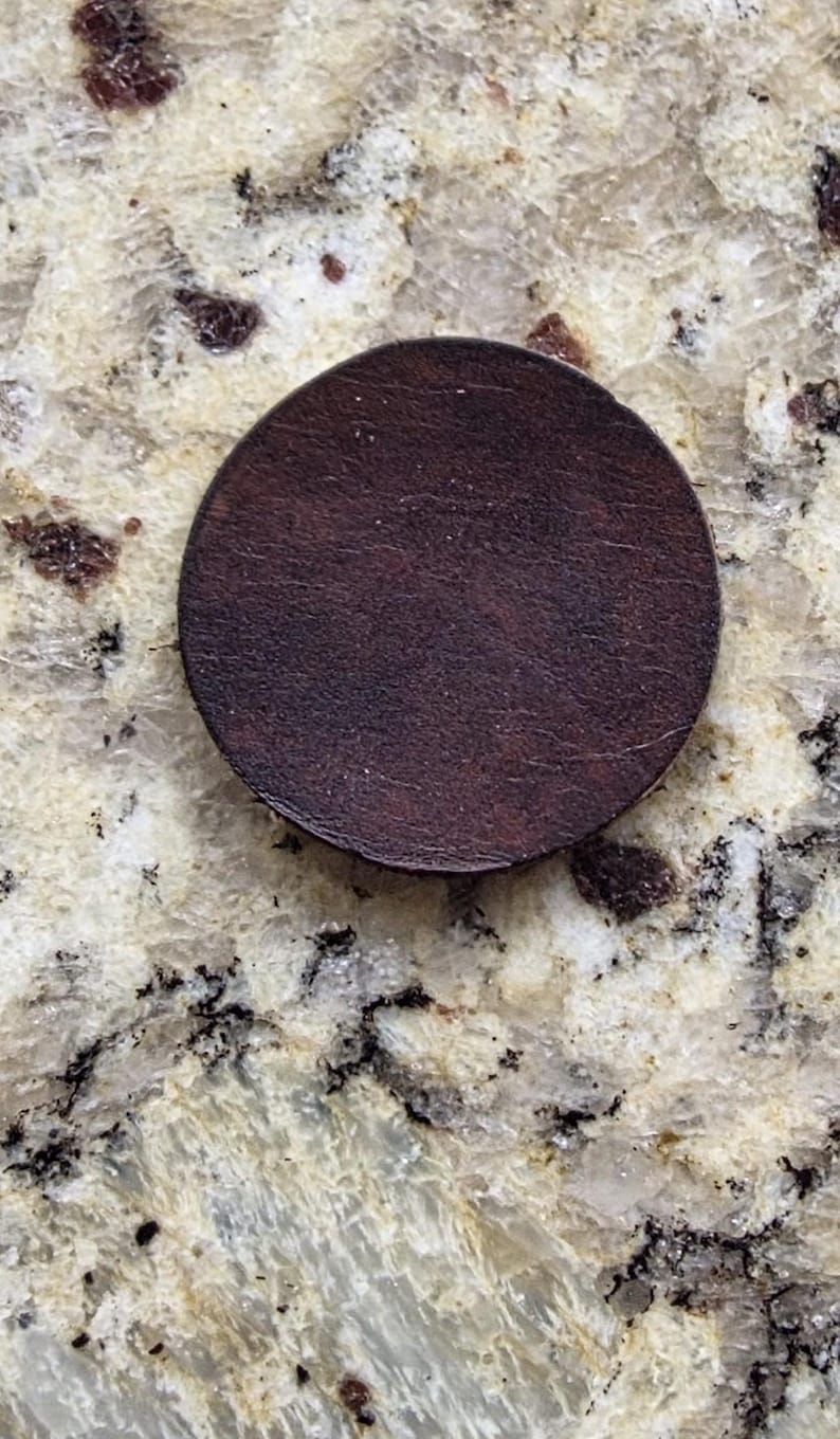 Peut inclure: Un dessous de verre circulaire en cuir marron fonc&eacute;. Le dessous de verre est de couleur unie avec une surface l&eacute;g&egrave;rement textur&eacute;e. Le dessous de verre est pos&eacute; sur un comptoir en granit clair et mouchet&eacute;.