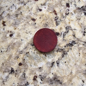 Peut inclure: Un petit disque rond en bois rouge fonc&eacute;. Le disque est centr&eacute; dans l'image et semble &ecirc;tre fait d'une seule pi&egrave;ce de bois. L'arri&egrave;re-plan est une surface claire et mouchet&eacute;e.