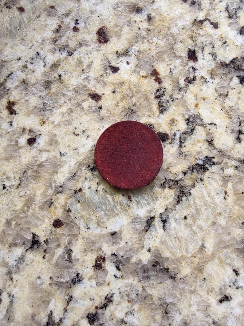 Peut inclure: Un petit disque rond en bois rouge fonc&eacute;. Le disque est centr&eacute; dans l'image et semble &ecirc;tre fait d'une seule pi&egrave;ce de bois. L'arri&egrave;re-plan est une surface claire et mouchet&eacute;e.