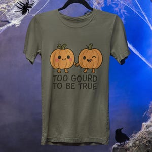 Puede incluir: Camiseta verde oliva con un gráfico de dos calabazas sonrientes tomadas de la mano. El texto "TOO GOURD TO BE TRUE" está impreso debajo. La camiseta está colgada en una percha negra.