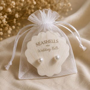 May include: A pair of white pearl stud earrings displayed on a cream-colored, shell-shaped card with the text SEASHELLS AND Wedding Bells printed in gold. The card is inside a small, sheer white organza drawstring pouch with a ribbon closure.