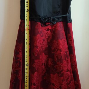 May include: A formal dress featuring a red floral lace overlay. The dress has a black satin waistband and a layered skirt. The total length of the dress is approximately 44 inches.