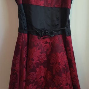 May include: A formal dress with a burgundy floral pattern over a black underlay. The dress features a black satin waist band and a high-low hemline. The dress is suspended by thin black straps.