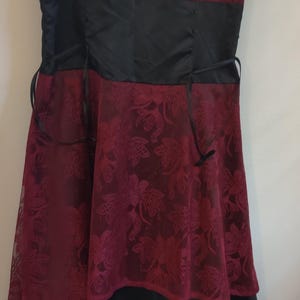 May include: Burgundy-colored dress with a floral lace overlay. The bodice has a black satin band and spaghetti straps. The skirt features a layered design with a black satin hem. A formal dress.