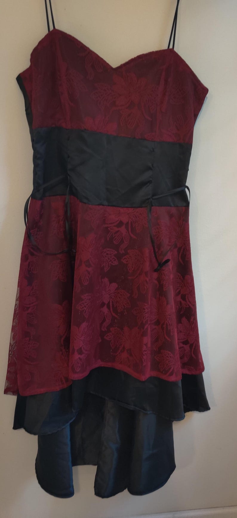 May include: Burgundy-colored dress with a floral lace overlay. The bodice has a black satin band and spaghetti straps. The skirt features a layered design with a black satin hem. A formal dress.