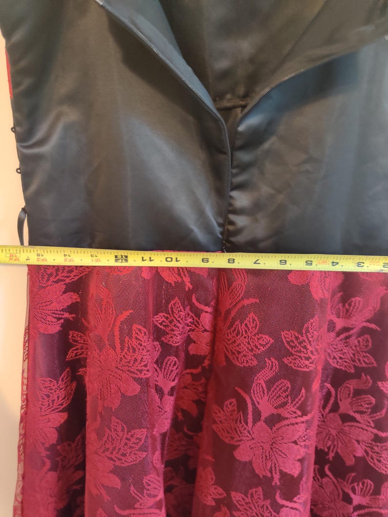 May include: A black satin dress bodice with a zipper closure, transitioning to a burgundy floral lace skirt. The dress is measured with a yellow tape measure. The skirt has a floral pattern.