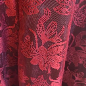 May include: Close-up of a fabric with a floral pattern. The fabric is a deep burgundy color with a repeating design of red flowers and leaves. The texture appears to be woven, with a slight sheen. The folds of the fabric create shadows, adding depth to the image.