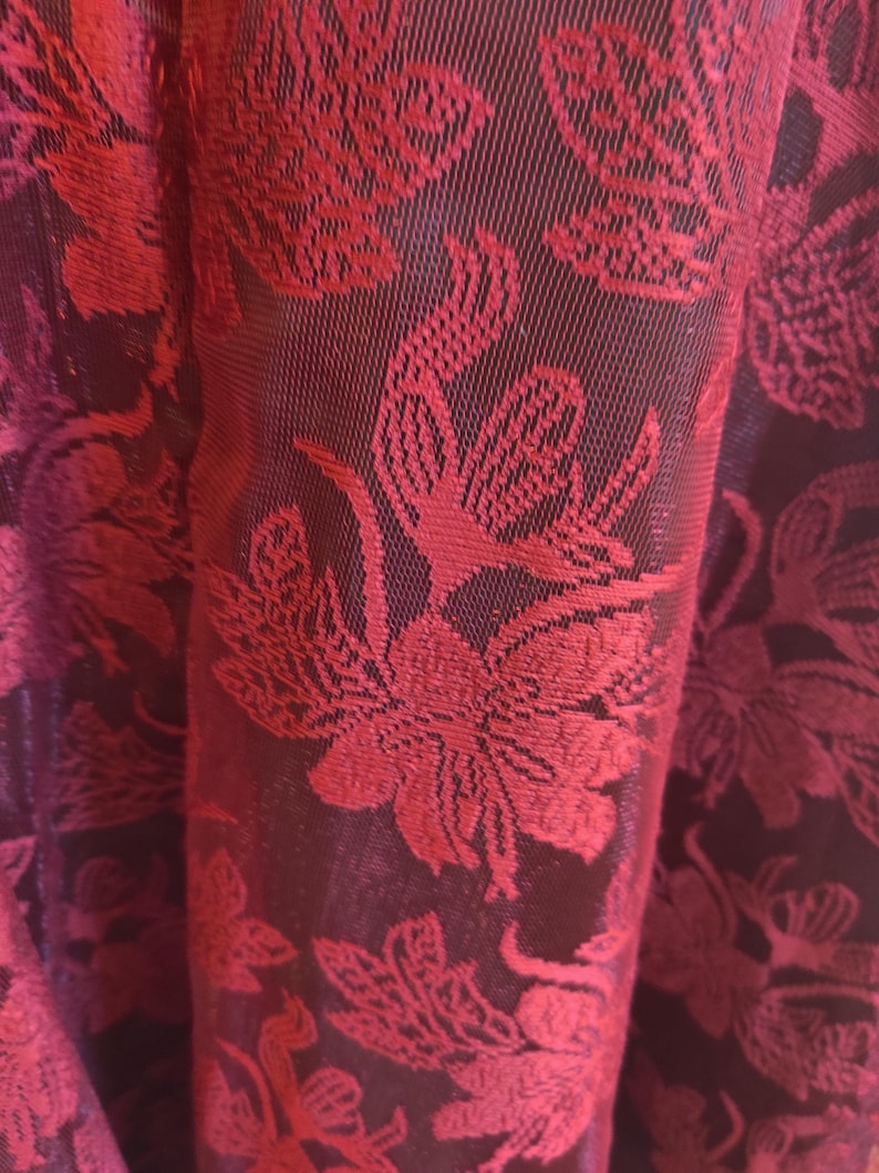 May include: Close-up of a fabric with a floral pattern. The fabric is a deep burgundy color with a repeating design of red flowers and leaves. The texture appears to be woven, with a slight sheen. The folds of the fabric create shadows, adding depth to the image.