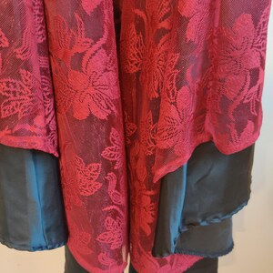 May include: Close-up of a layered garment featuring a sheer, burgundy floral lace overlay. The garment has a dark teal layer and a black layer underneath, creating a contrast in textures and colors. The floral pattern is detailed.
