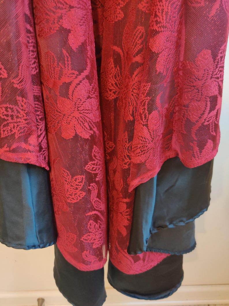 May include: Close-up of a layered garment featuring a sheer, burgundy floral lace overlay. The garment has a dark teal layer and a black layer underneath, creating a contrast in textures and colors. The floral pattern is detailed.