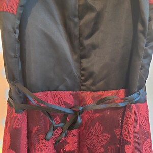 May include: Close-up of a formal dress featuring a black bodice and a burgundy skirt with a floral pattern. A black ribbon belt is tied at the waist, adding a defined shape to the dress. The dress has a satin-like texture.