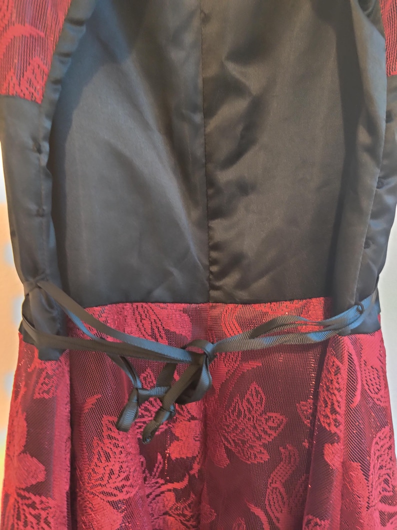 May include: Close-up of a formal dress featuring a black bodice and a burgundy skirt with a floral pattern. A black ribbon belt is tied at the waist, adding a defined shape to the dress. The dress has a satin-like texture.